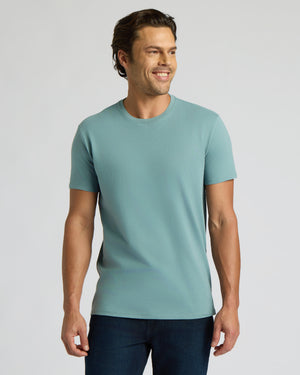 A man wearing a light teal short-sleeve crewneck t-shirt and dark blue jeans, standing against a solid light gray background.