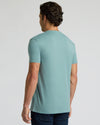 A man viewed from the back wearing a light blue-green crewneck t-shirt and dark blue jeans, standing against a plain light gray studio background.