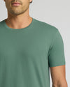 A close-up of a man wearing a muted sage green crew-neck t-shirt against a light gray background.