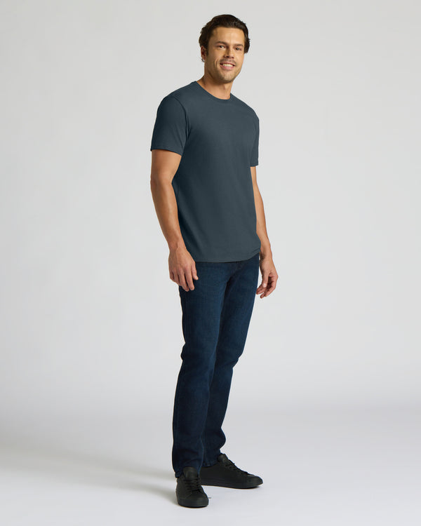 A man modeling a dark slate blue crew-neck t-shirt, dark blue jeans, and black sneakers against a light gray studio background.
