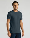 A man wearing a dark slate blue crewneck t-shirt and dark blue jeans, standing against a light gray studio background.