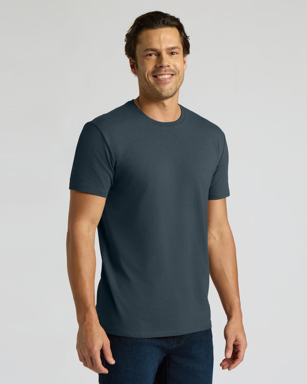 A man wearing a dark slate blue crewneck t-shirt and dark blue jeans, standing against a light gray studio background.