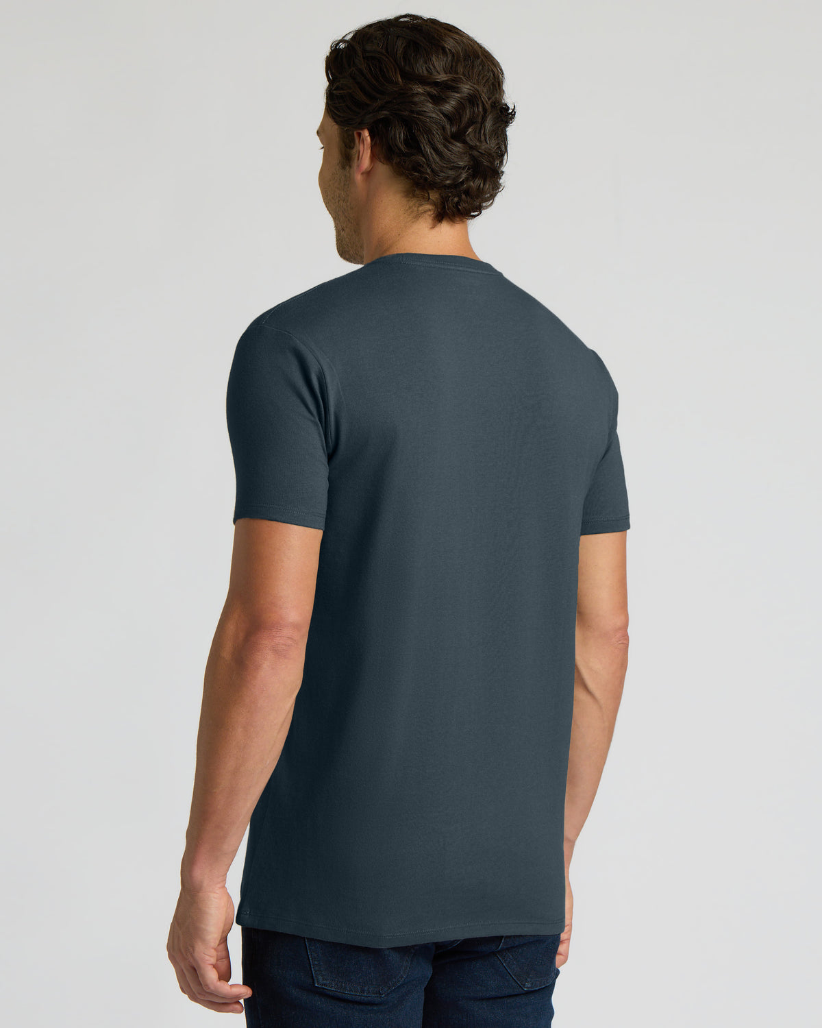 A man viewed from the back wearing a dark slate blue crewneck t-shirt and dark jeans, set against a plain light gray studio background.