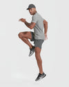 Man wearing an Iron Gray Active Crew Neck Tee, gray athletic shorts, black running shoes, and a gray cap, performing a high knee exercise against a plain white background.