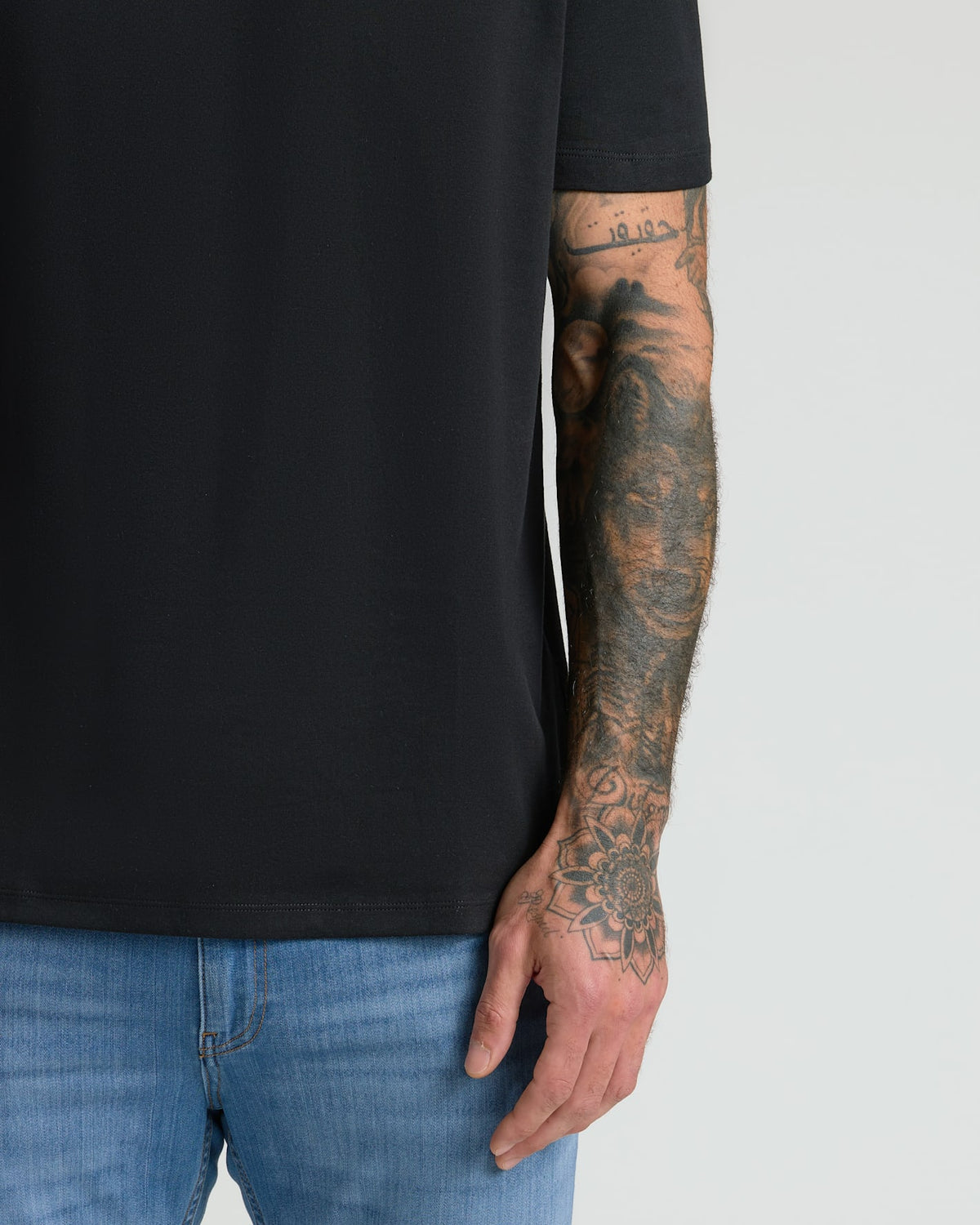 Close-up of a person wearing a black crew neck t-shirt from the All Black Pima Cotton Blend Crew 7-Pack paired with blue jeans, showcasing a tattooed arm.
