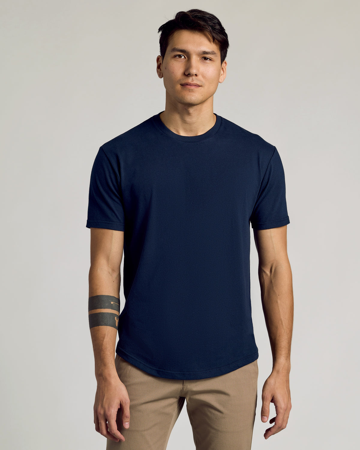 Man wearing a navy Classic Curved Hem Crew Neck 3-Pack t-shirt with short sleeves and a curved hem, styled with tan pants against a neutral background.
