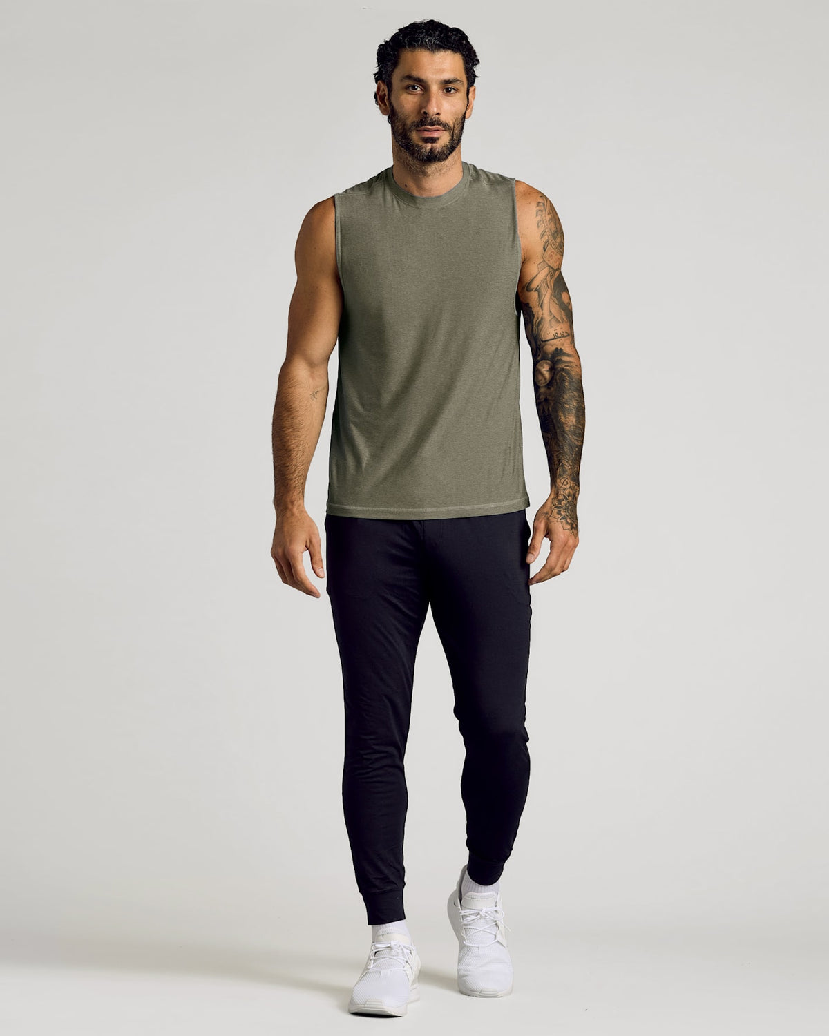 Man wearing a Heather Military Green Sleeveless Active Muscle Tee paired with black joggers and white sneakers, showcasing a casual and athletic men's basics look.