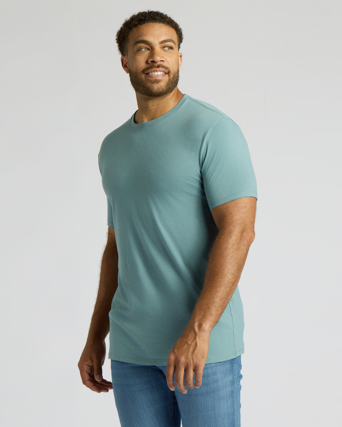 A man wearing a fitted short-sleeve crewneck t-shirt in a muted teal-blue color, paired with blue jeans, standing against a light gray studio background.