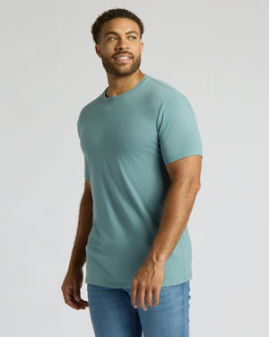 A man wearing a fitted short-sleeve crewneck t-shirt in a muted teal-blue color, paired with blue jeans, standing against a light gray studio background.