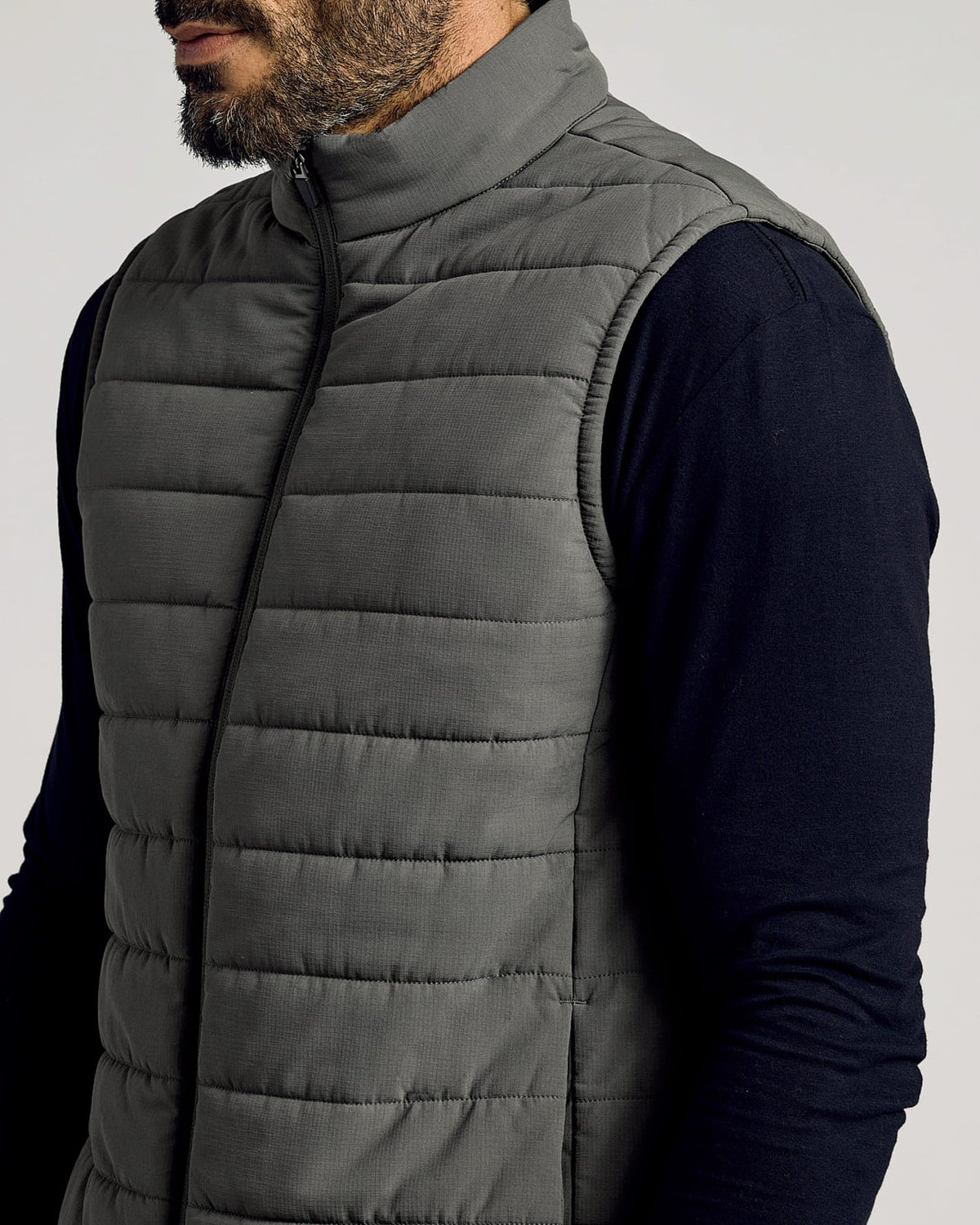 Man wearing a gray Carbon Puffer Vest over a black long-sleeve shirt, featuring a high collar and horizontal quilted stitching.