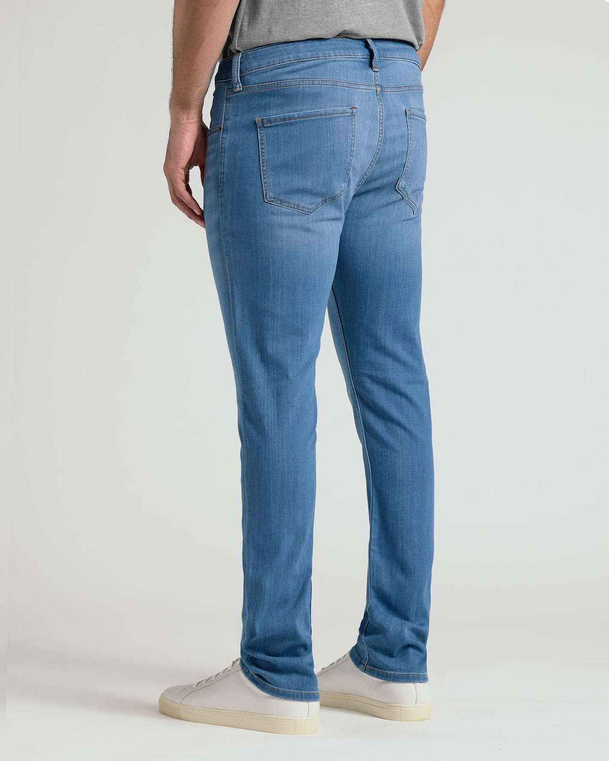 Rear view of a person wearing slim-fit blue jeans with white sneakers against a plain light gray studio background.