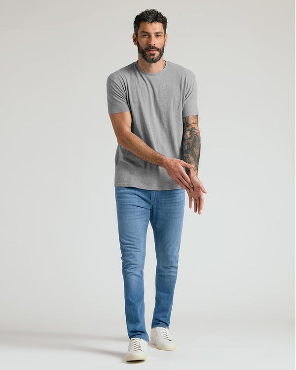 A man modeling a light gray crew-neck t-shirt and blue slim-fit jeans with white sneakers, standing against a light gray studio background.
