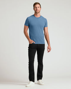 True ClassicA man modeling a short-sleeve crewneck t-shirt in a medium blue color, paired with black pants and white sneakers, standing against a light gray studio background.