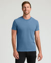 A smiling man wearing a solid medium blue crew-neck t-shirt and black pants, standing against a light gray studio background.