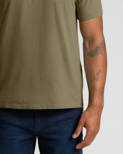 True ClassicA close-up of a person wearing an olive green short-sleeve t-shirt and dark blue jeans, showing the side of the torso and arm with a tattoo against a light gray background.