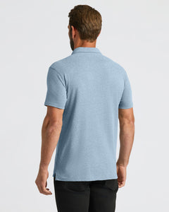 True ClassicA man viewed from the back wearing a light heather blue short-sleeve polo shirt and black pants, against a plain light gray studio background.