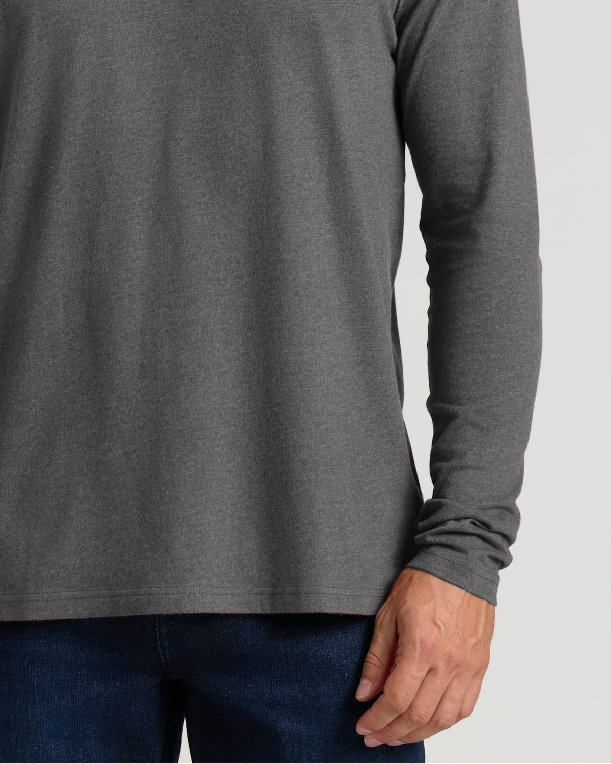 A close-up of a person wearing a dark heather gray long-sleeve shirt with dark blue jeans, cropped from the chest down against a light gray background.
