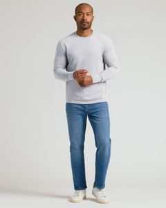 True ClassicA man wearing a light gray long-sleeve crewneck sweater, blue jeans, and white sneakers, standing against a plain light gray studio background.