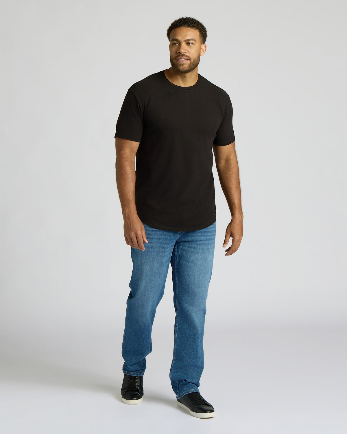 A man wearing a Black Tall Heavyweight Curved Hem Crew shirt paired with blue jeans and black sneakers, standing on a plain light background.