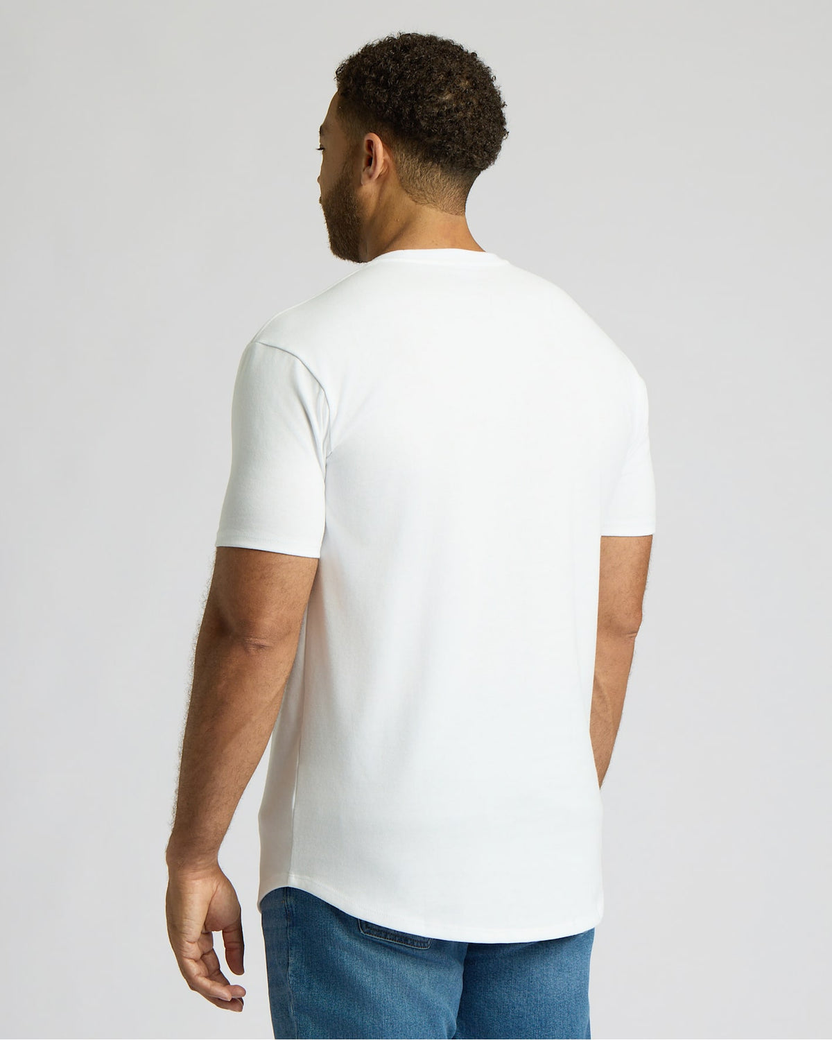 Man wearing an all white crew neck t-shirt from the All White Tall Heavyweight Curved Hem Crew 3-Pack, paired with blue jeans, shown from the back in a casual pose.