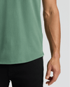 Close-up of a man's arm beside a green Neo Fern Heavyweight Curved Hem Henley shirt paired with black pants, showcasing the curved hem detail of this men's basics top.