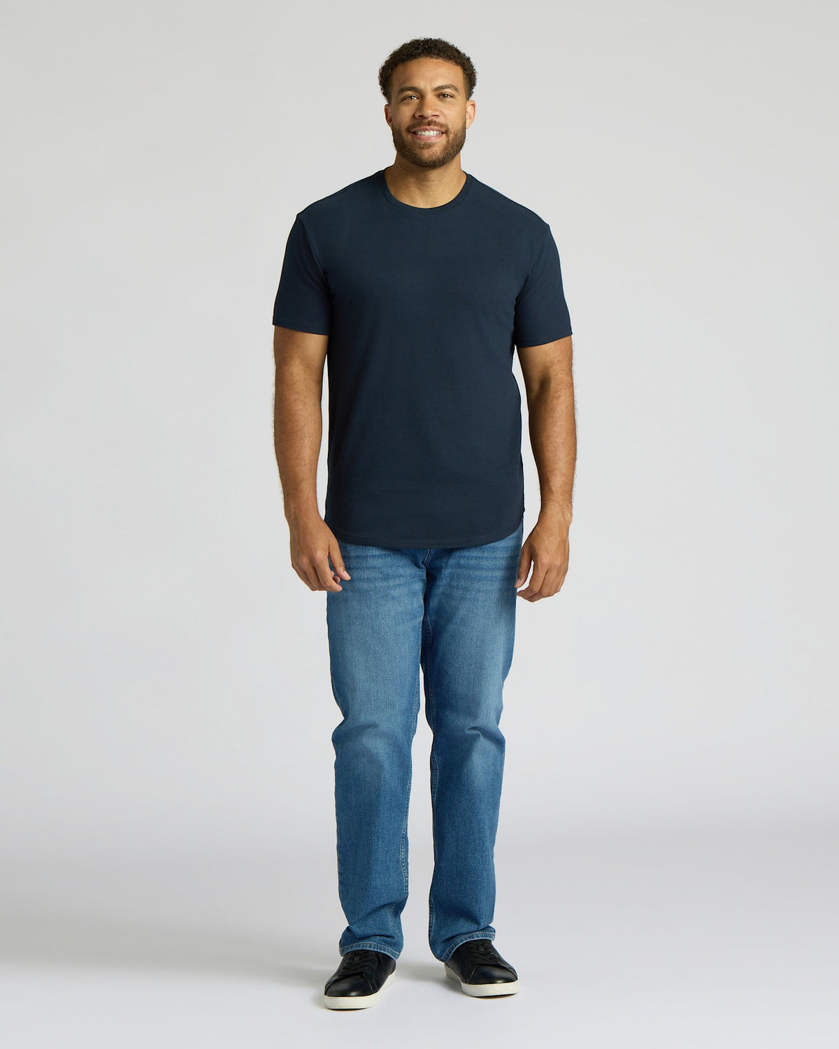 Man wearing a navy tall heavyweight curved hem crew shirt paired with blue jeans and black sneakers, standing against a plain light gray background.