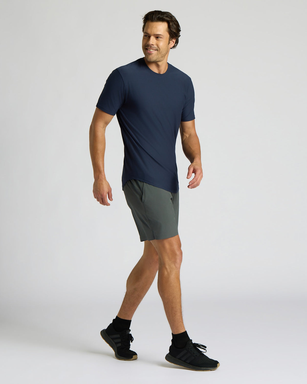 Man wearing a navy blue Active Curved Hem Crew 6-Pack t-shirt paired with gray athletic shorts and black sneakers, walking confidently against a plain background.
