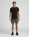 Man wearing an All Black Active Curved Hem Crew 6-Pack black short sleeve crew neck t-shirt paired with gray athletic shorts and black sneakers, standing against a plain background.