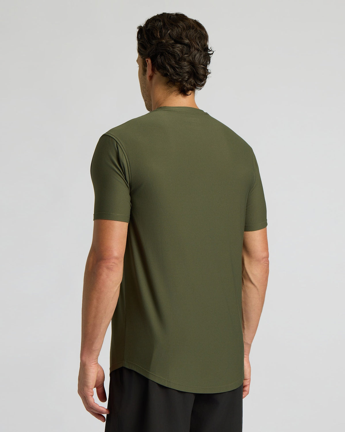 A man viewed from the back wearing a short-sleeve olive green athletic t-shirt and black pants, set against a plain light gray studio background.