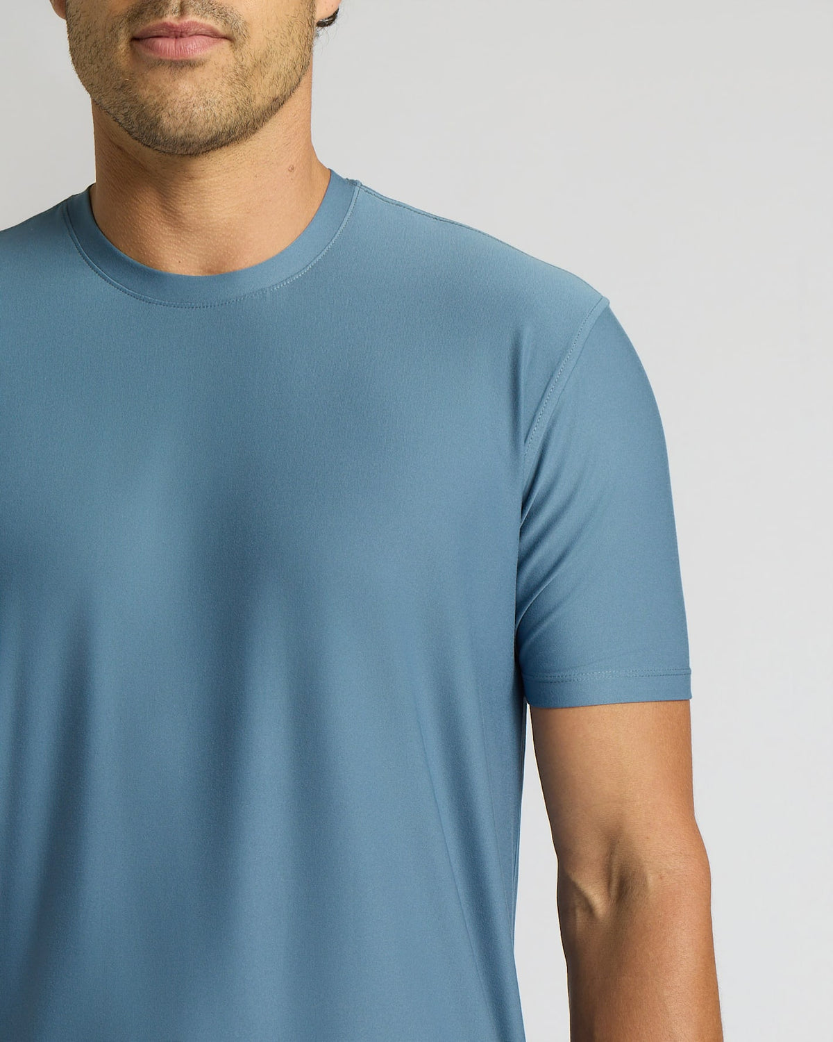 A close-up of a man wearing a short-sleeve crew-neck t-shirt in a dusty slate blue color, shown from the chin down against a light gray studio background.