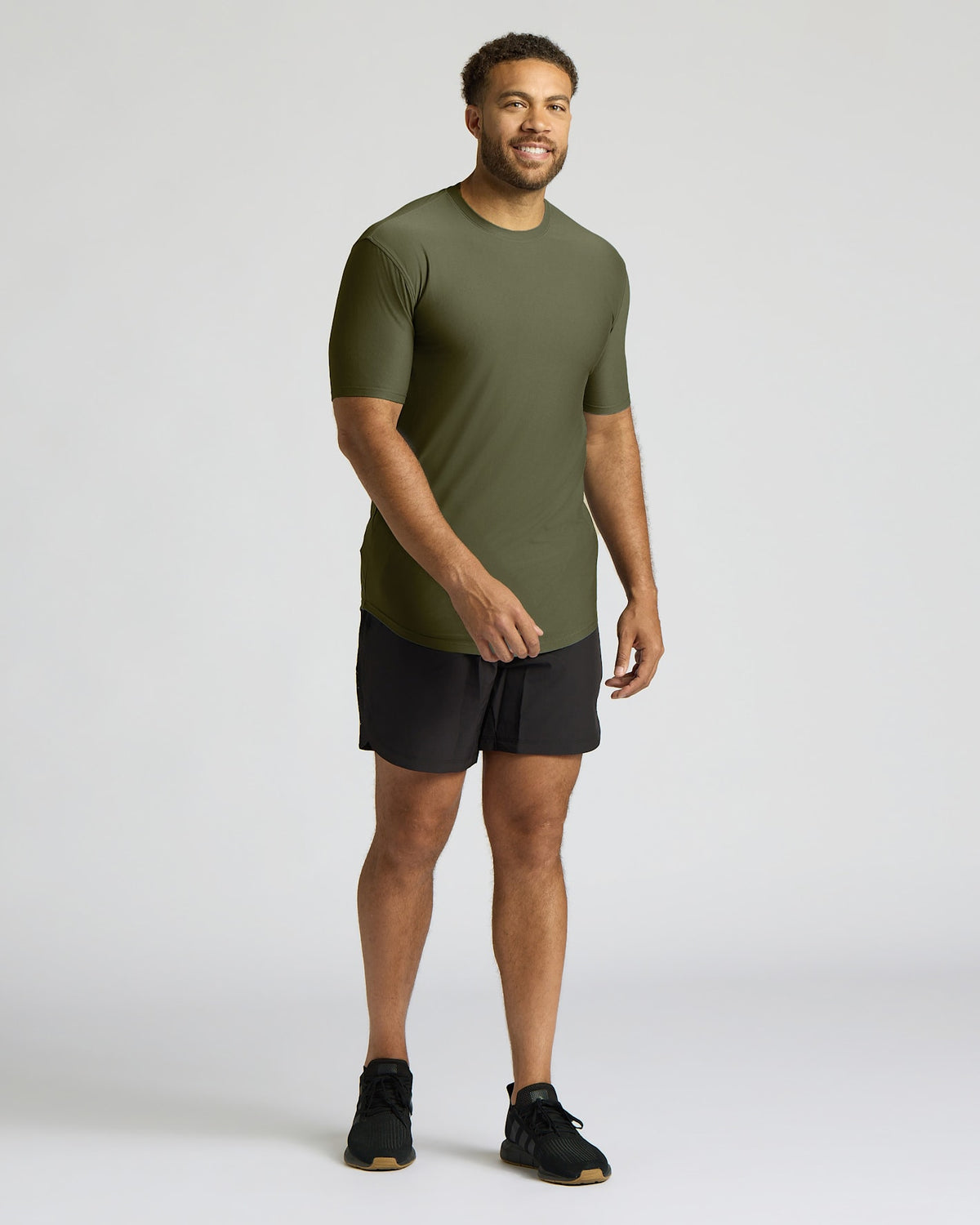 A man modeling a short-sleeve athletic t-shirt in olive green paired with black athletic shorts and black sneakers, standing against a light gray studio background.