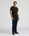 Man wearing a black Heavyweight Curved Hem Crew 6-Pack t-shirt paired with dark blue jeans and black shoes, standing against a plain light background.