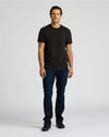 Male model wearing an All Black Heavyweight Curved Hem Crew 6-Pack t-shirt with dark jeans and black shoes, standing against a plain light background, showcasing men's basics from True Classic.