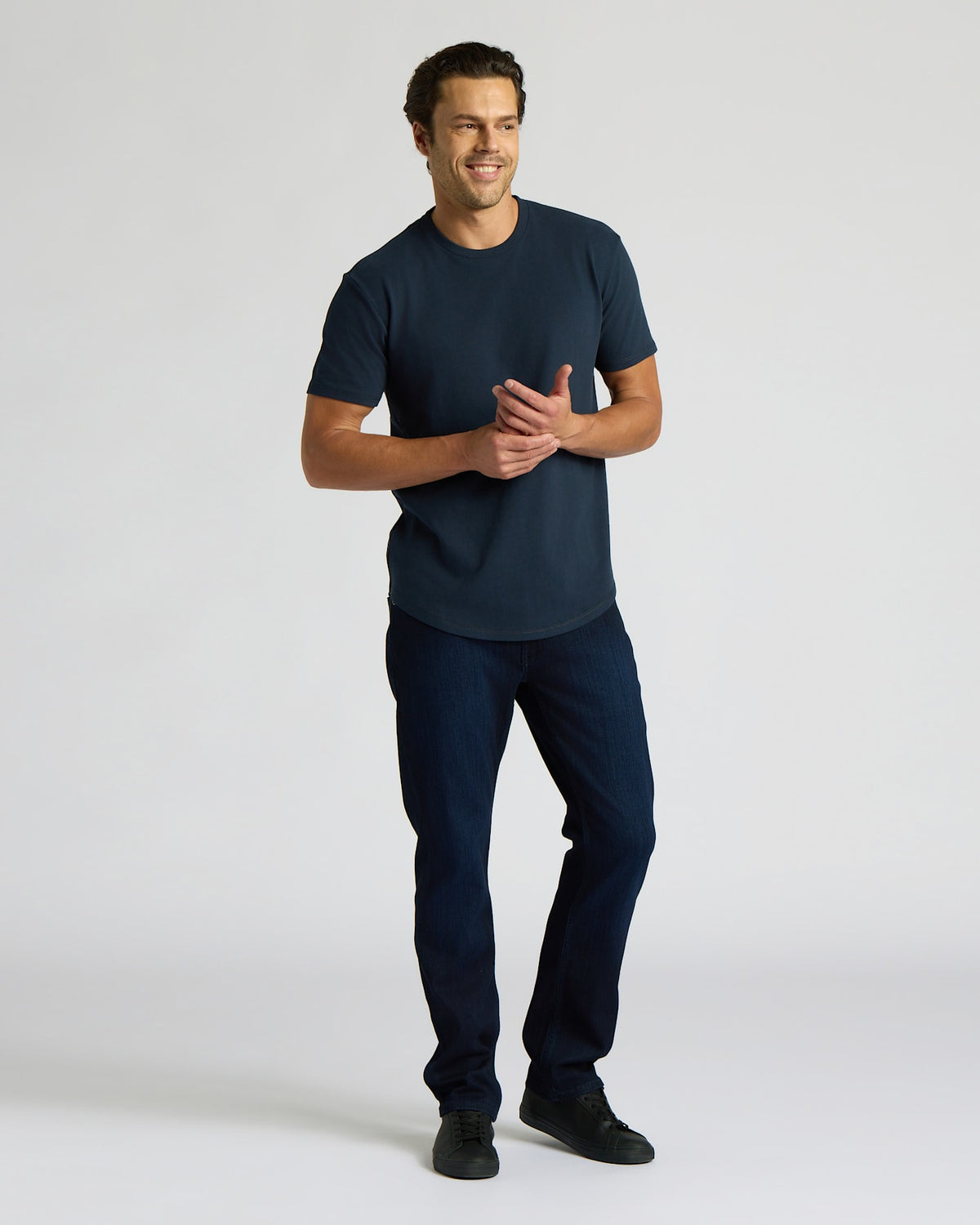 Man wearing a navy blue Heavyweight Curved Hem Crew 6-Pack t-shirt paired with dark jeans and black shoes, standing against a plain light background with a relaxed smile.