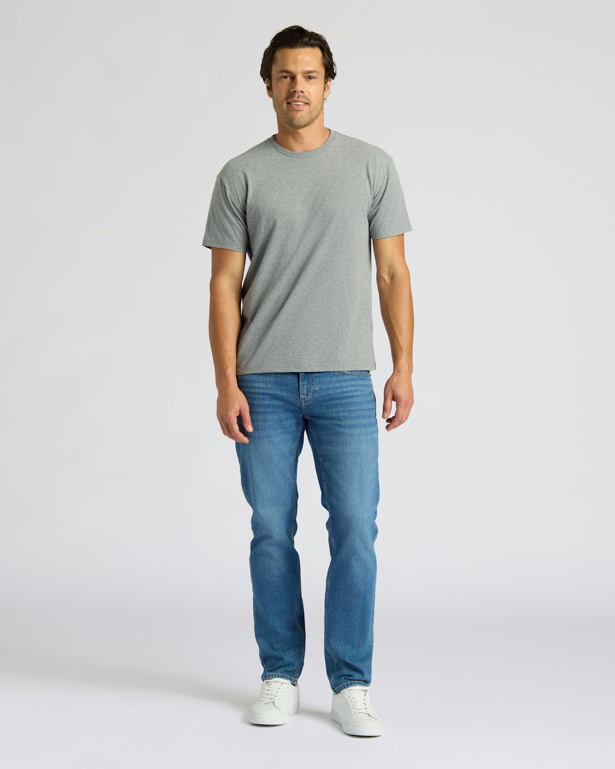 Man wearing a gray t-shirt and blue jeans on a white background. MODEL_INFO: Model is 6'0" wearing size L short