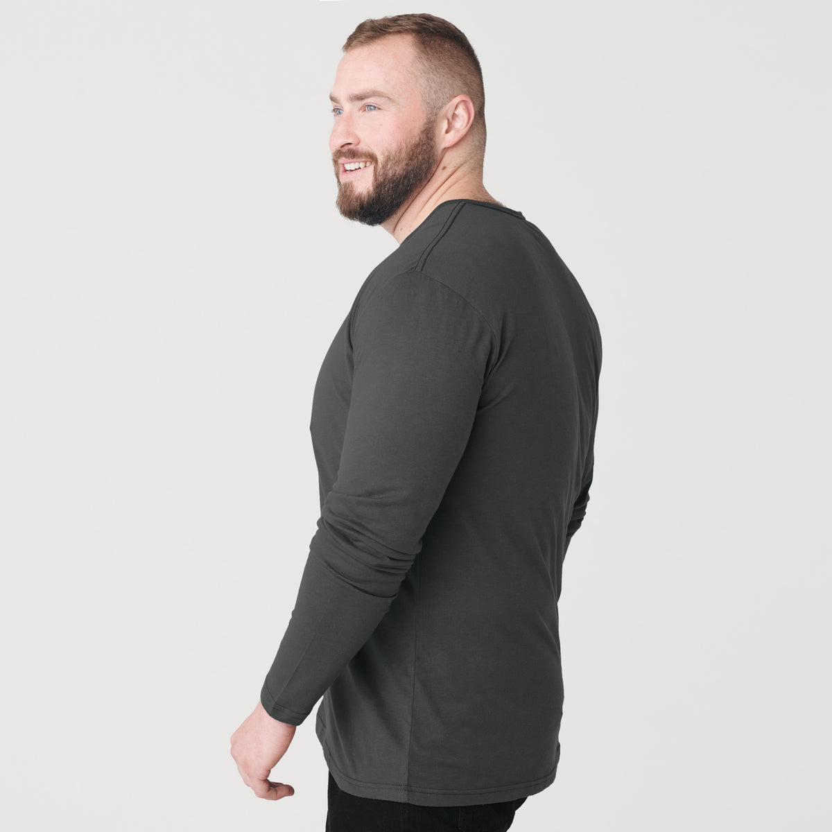 Man wearing Carbon Classic Long Sleeve Henley, dark pants, smiling slightly.